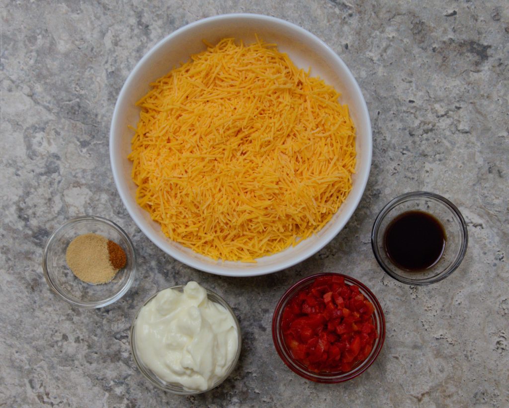 Ingredients for making pimento cheese. In each bowl there is onion powder, sharp cheddar cheese, worcestershire sauce, diced pimentos, and mayonnaise.  