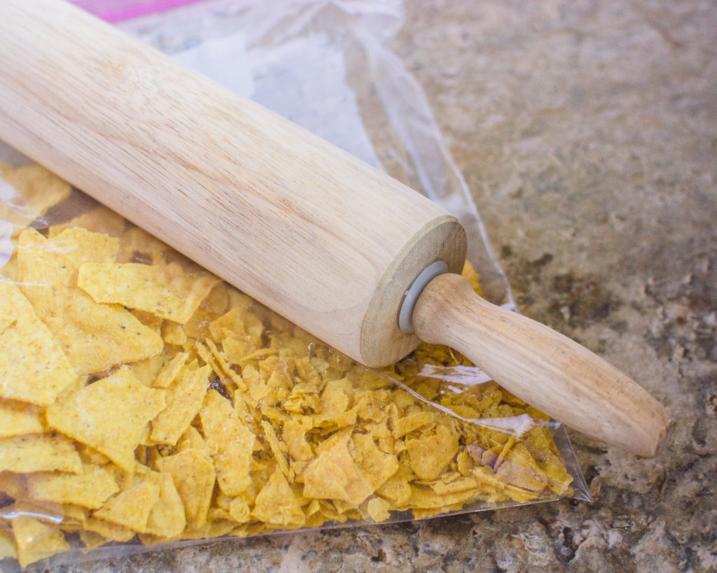 Tortilla chips in a freezer bag being crushed by a rolling pin that is rolling over the bag. 