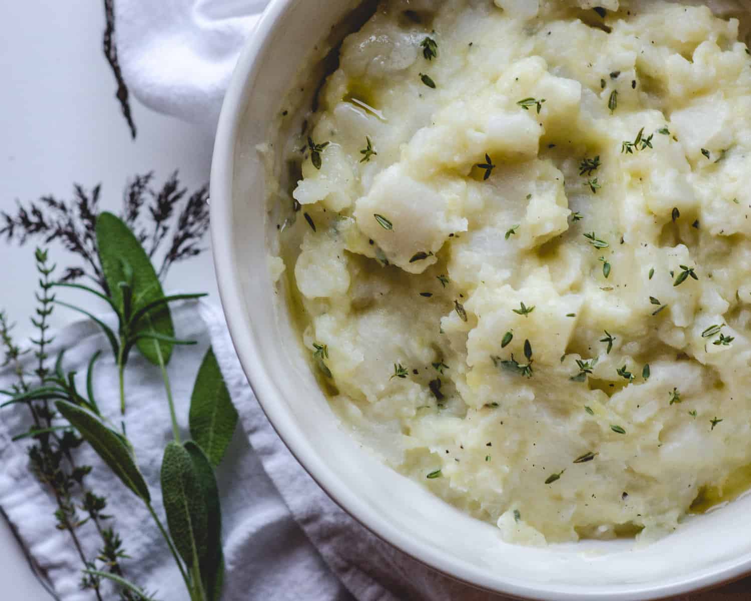 Bowl of mashed turnips and potatoes next to sage, thyme and rosemary sprigs. 
