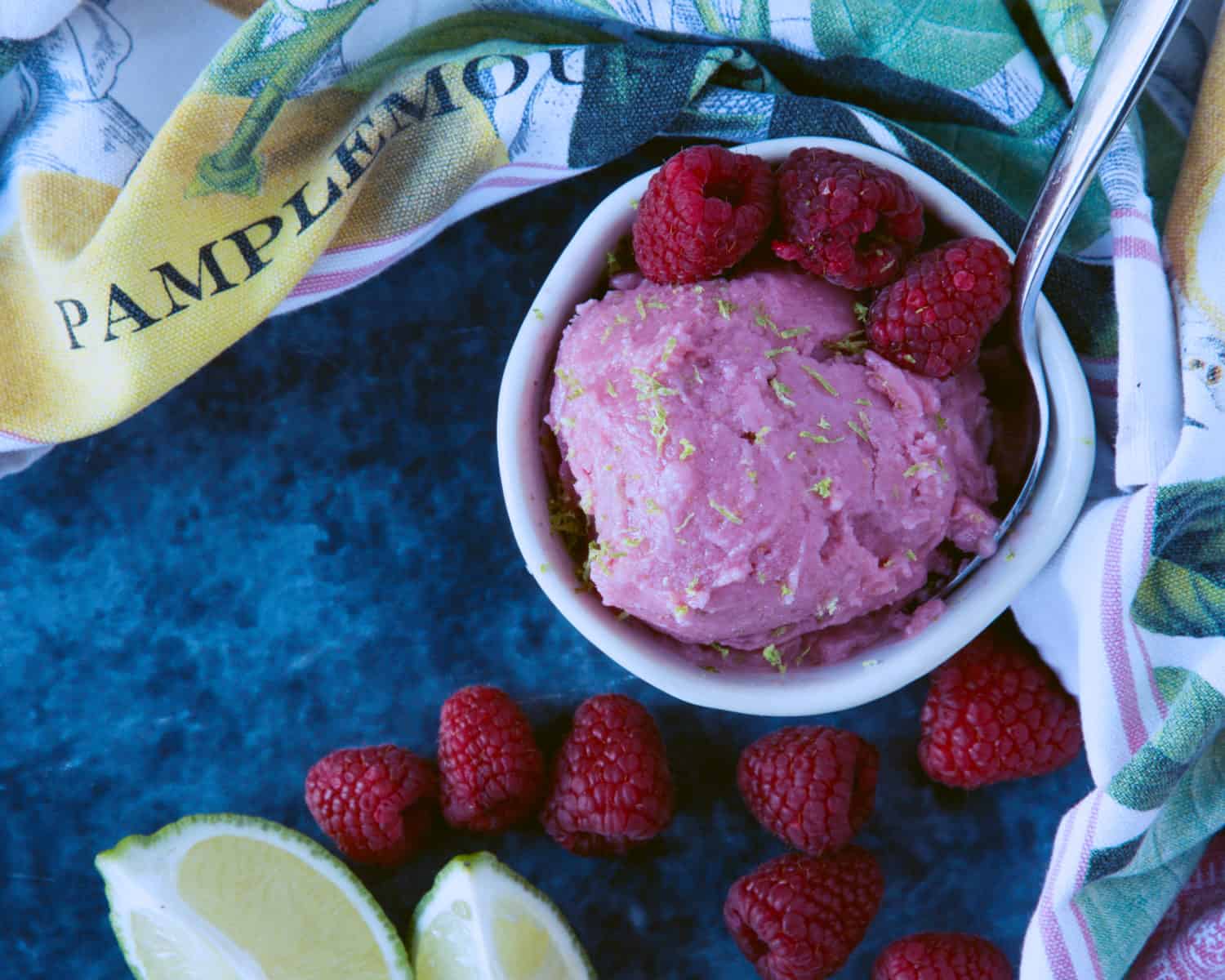 Overhead view of a bowl of raspberry sherbet with fresh berries, cut lime wedges and a hand towel. 