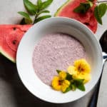 Bowl of watermelon chia seed pudding, spoon, flowers, mint sprigs and watermelon slices.