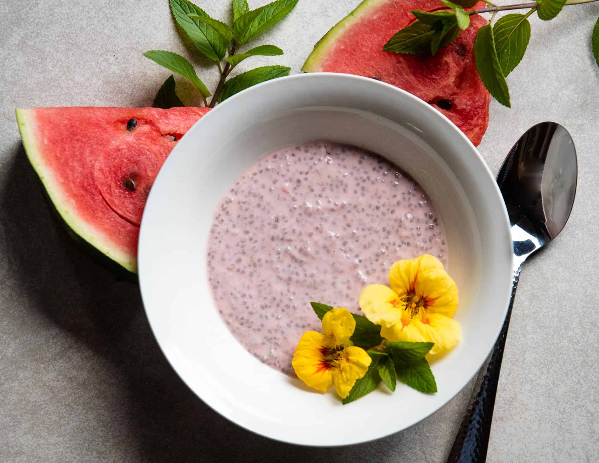 Bowl of watermelon chia seed pudding, spoon, flowers, mint sprigs and watermelon slices.