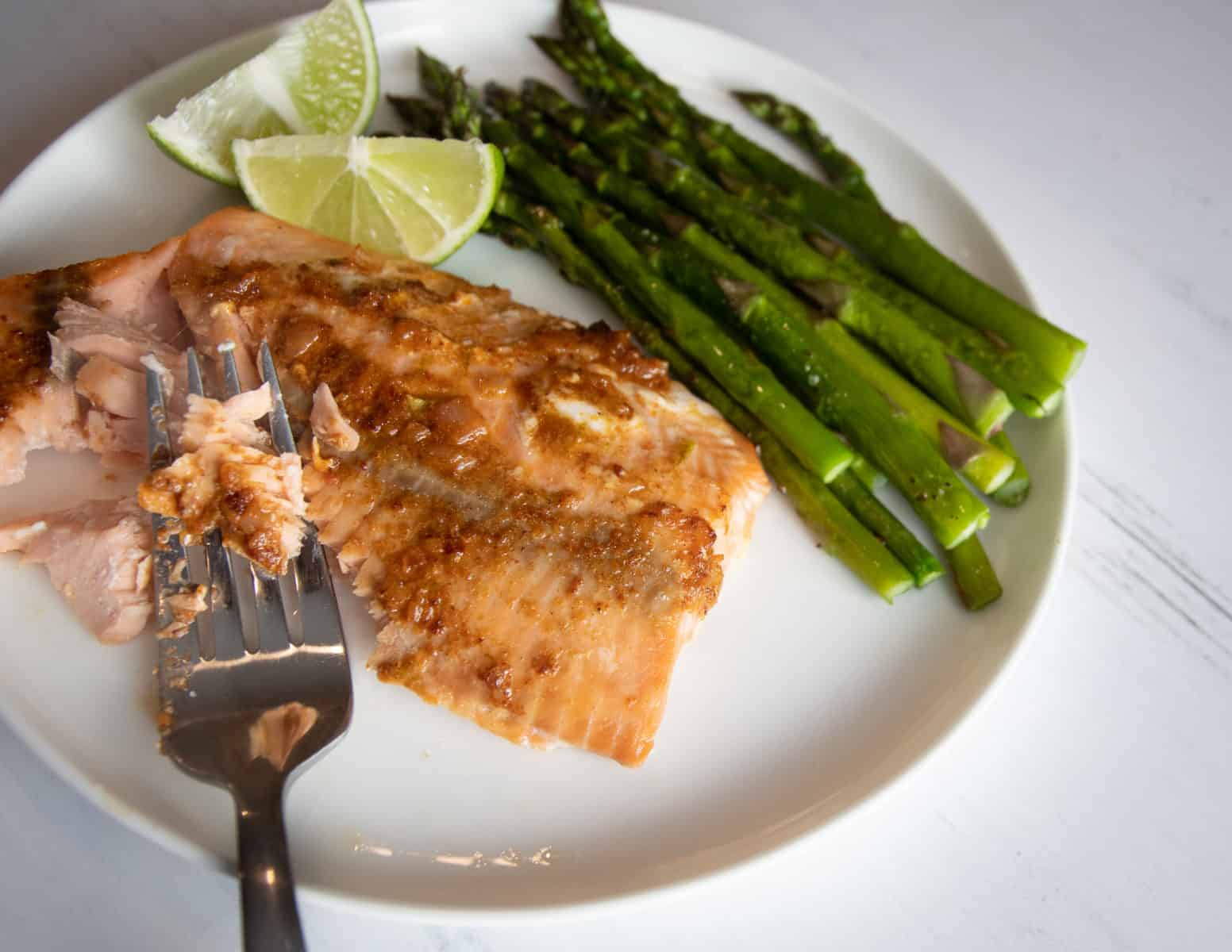 Plate of cooked salmon with roasted asparagus spears and two wedges of lime. Fork is flaking cooked salmon. 