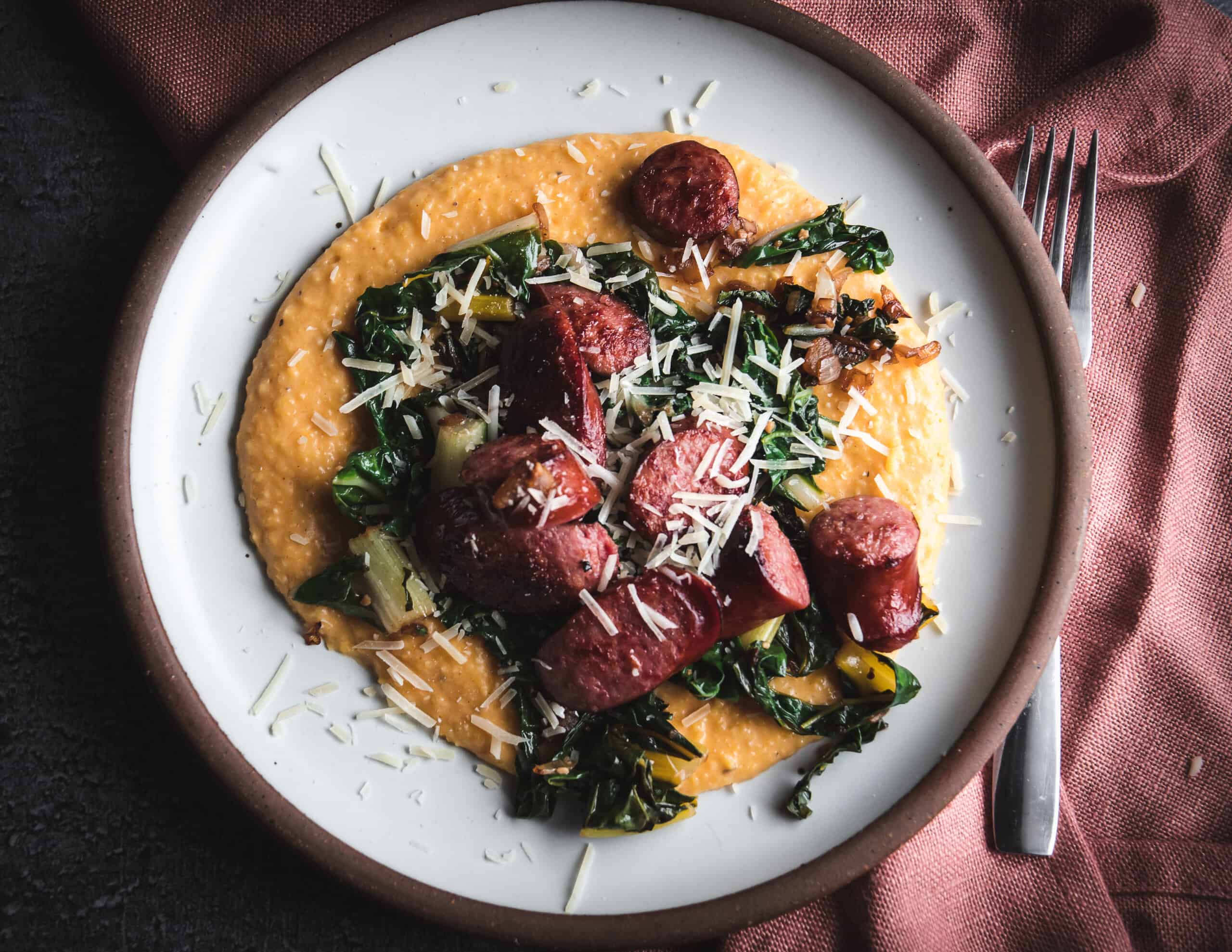 Plate of sweet potato polenta that is topped with cooked swiss chard, smoked sausage slices and grated parmesan cheese.