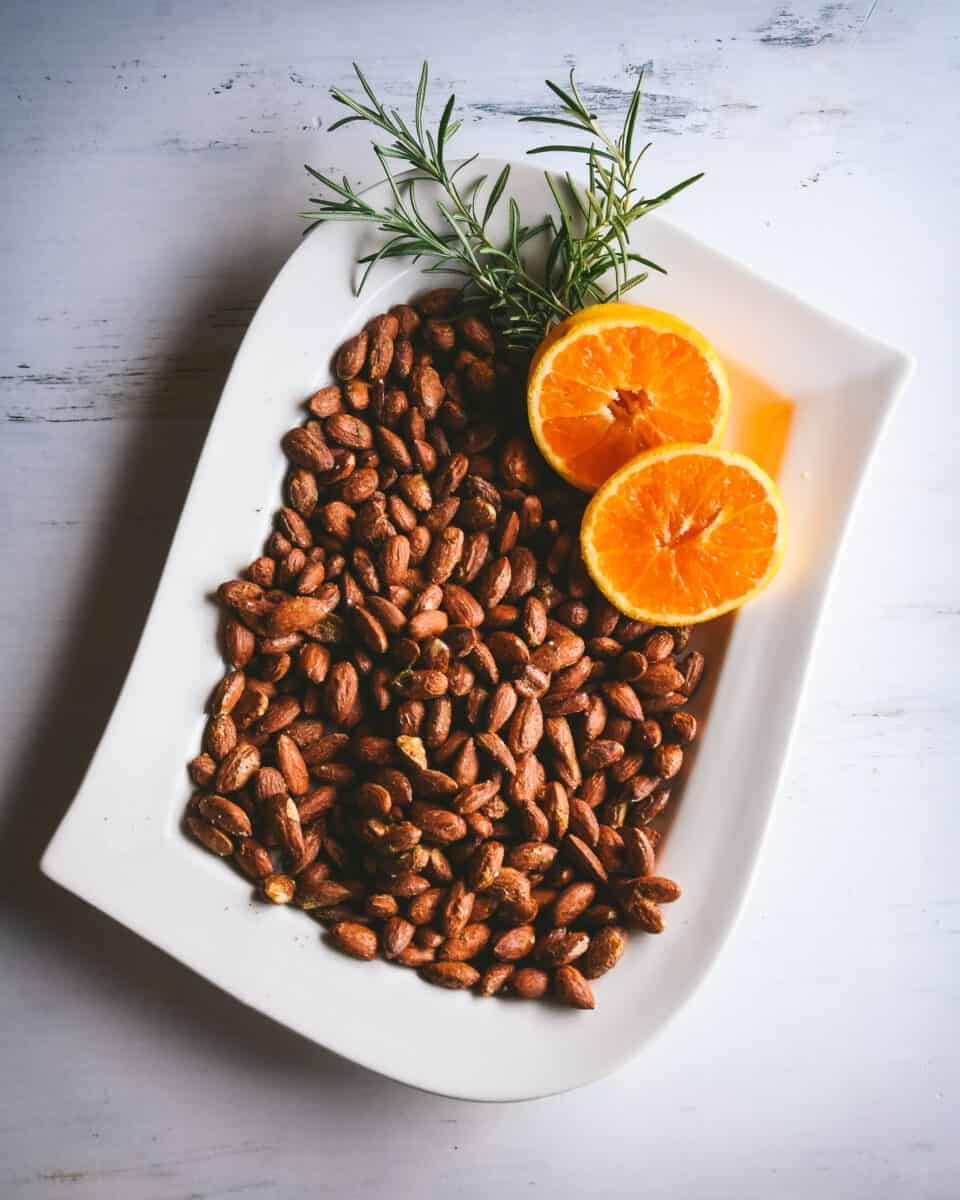 White platter of roasted almonds that are granished with two orange slices and a few sprigs of rosemary.