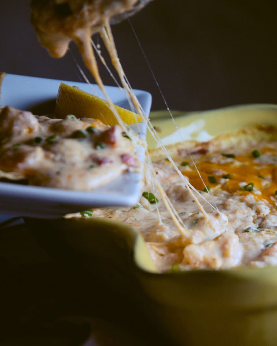 Shrimp dip being scooped out of casserole dish with melted cheese pulling across the plate.