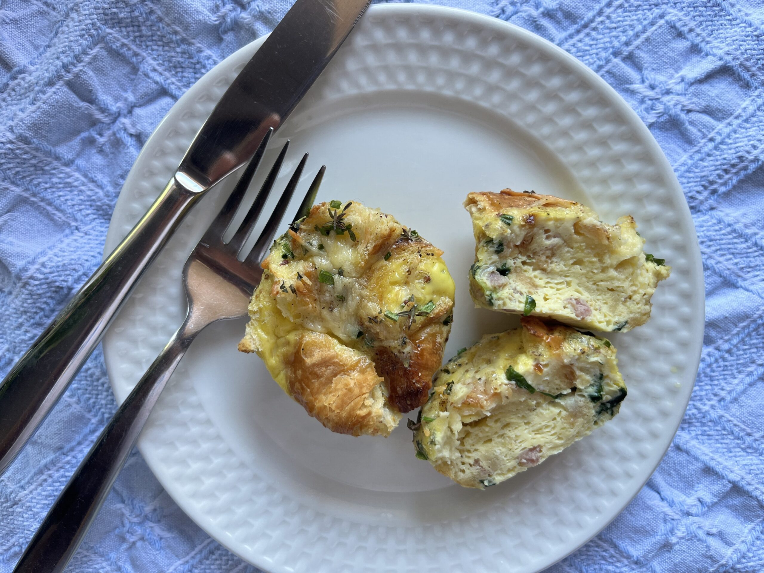 Plated savory croissant egg bites with a fork and knife. One egg bite is cut in half to show the fluffy, cooked interior.