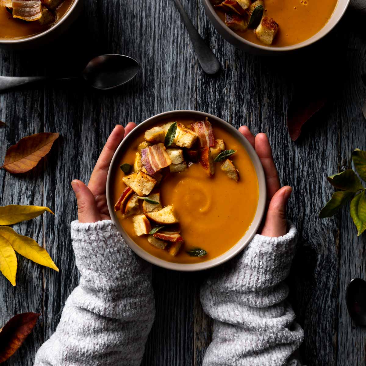 Pumpkin sweet potato soup in three bowls and garnished with croutons, sage, and cooked bacon.